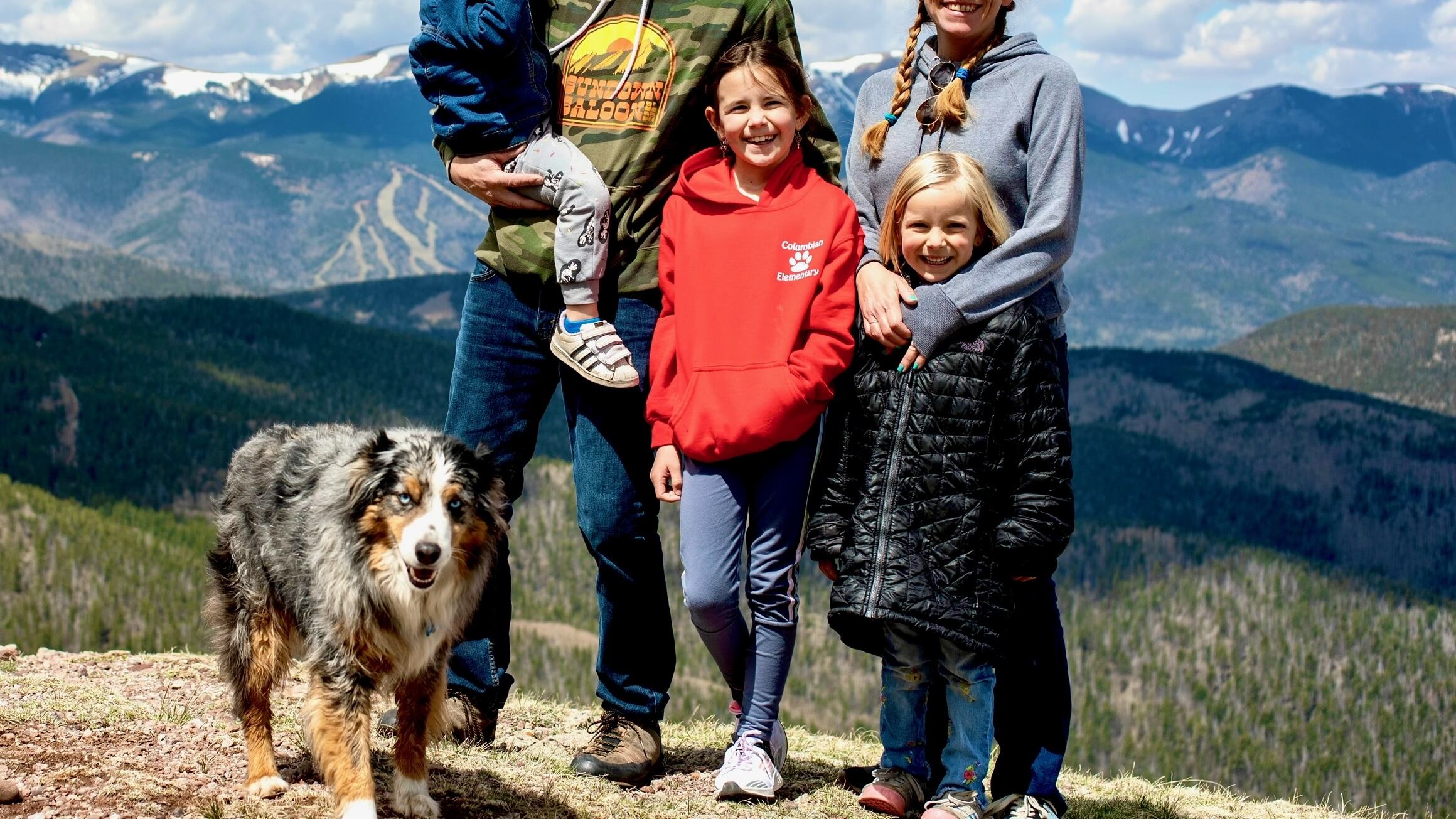 A dad, a mom, two daughters, and a son smile at the camera. They are outside with snow-capped mountains in the background.