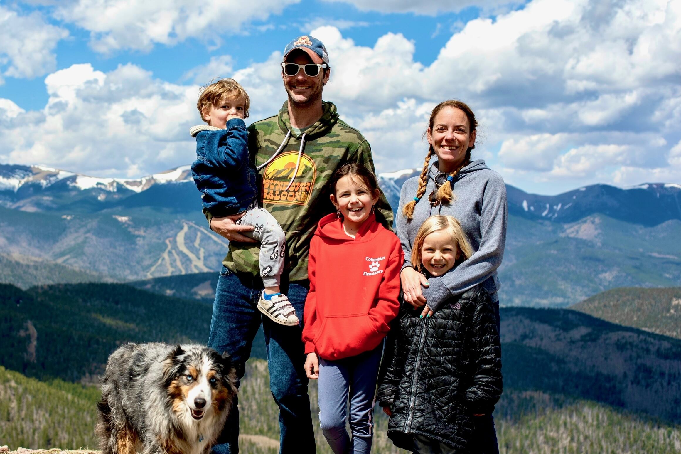 A dad, a mom, two daughters, and a son smile at the camera. They are outside with snow-capped mountains in the background.