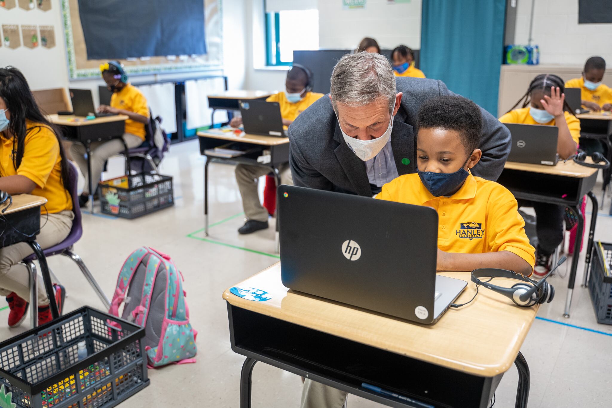 Governor Bill Lee, wearing a mask, looks over a student’s shoulder in a classroom, all the students wearing yellow shirts with protective masks.