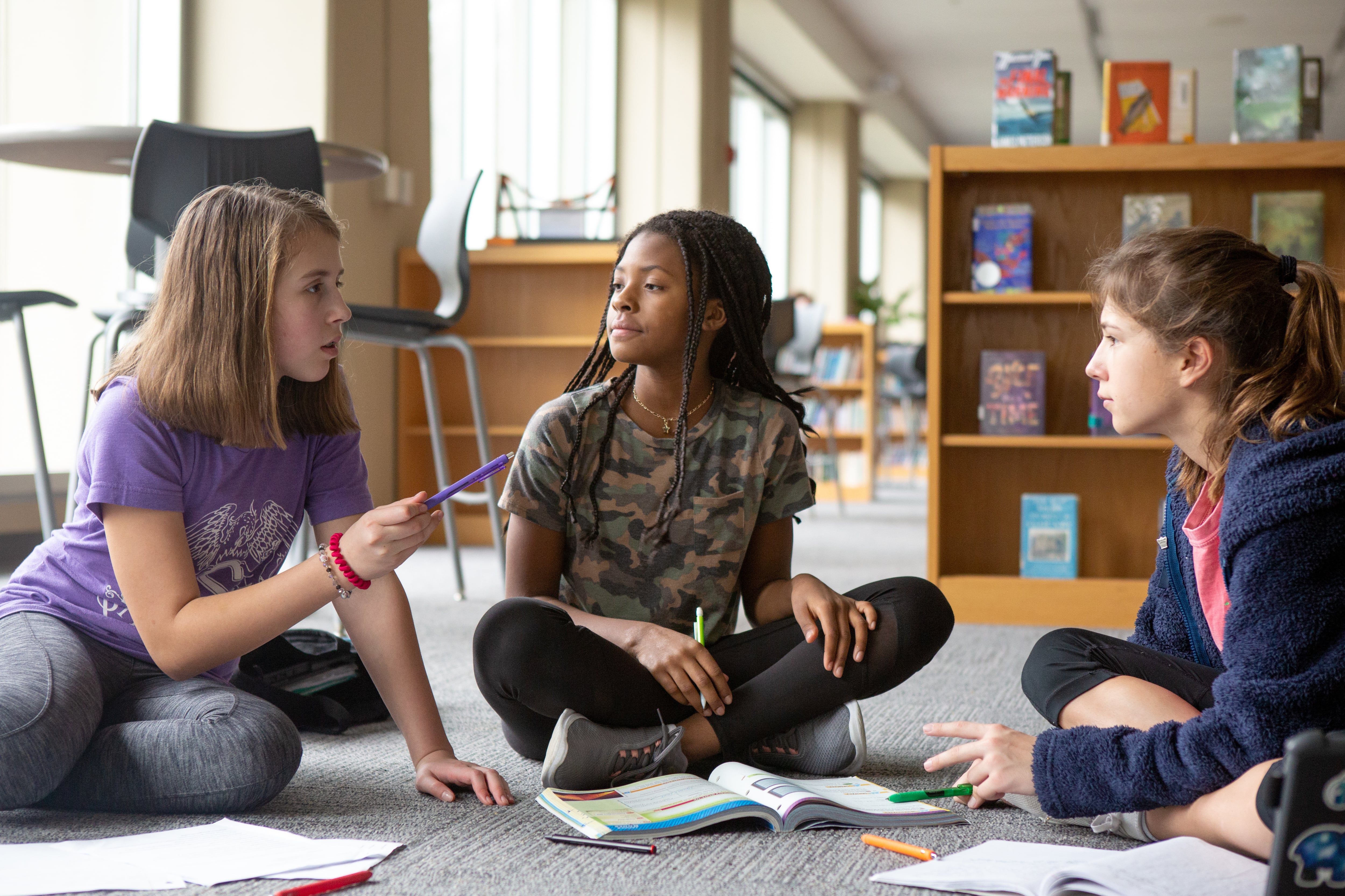 Three middle school students sit on the floor talking about school work with books and pencils on the ground in front of them.