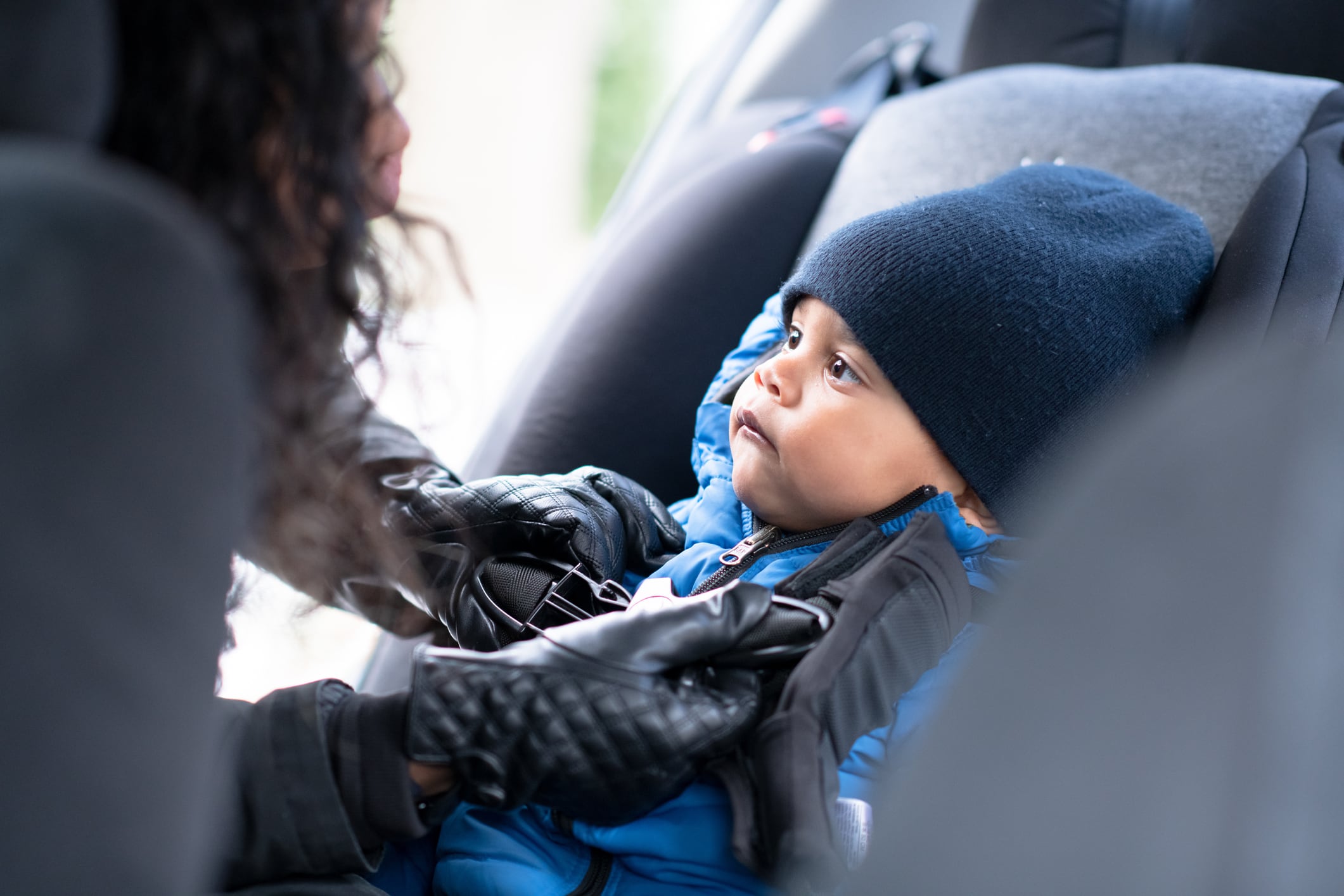A baby boy with a winter hat sits in in his car seat as his mother buckles him in.