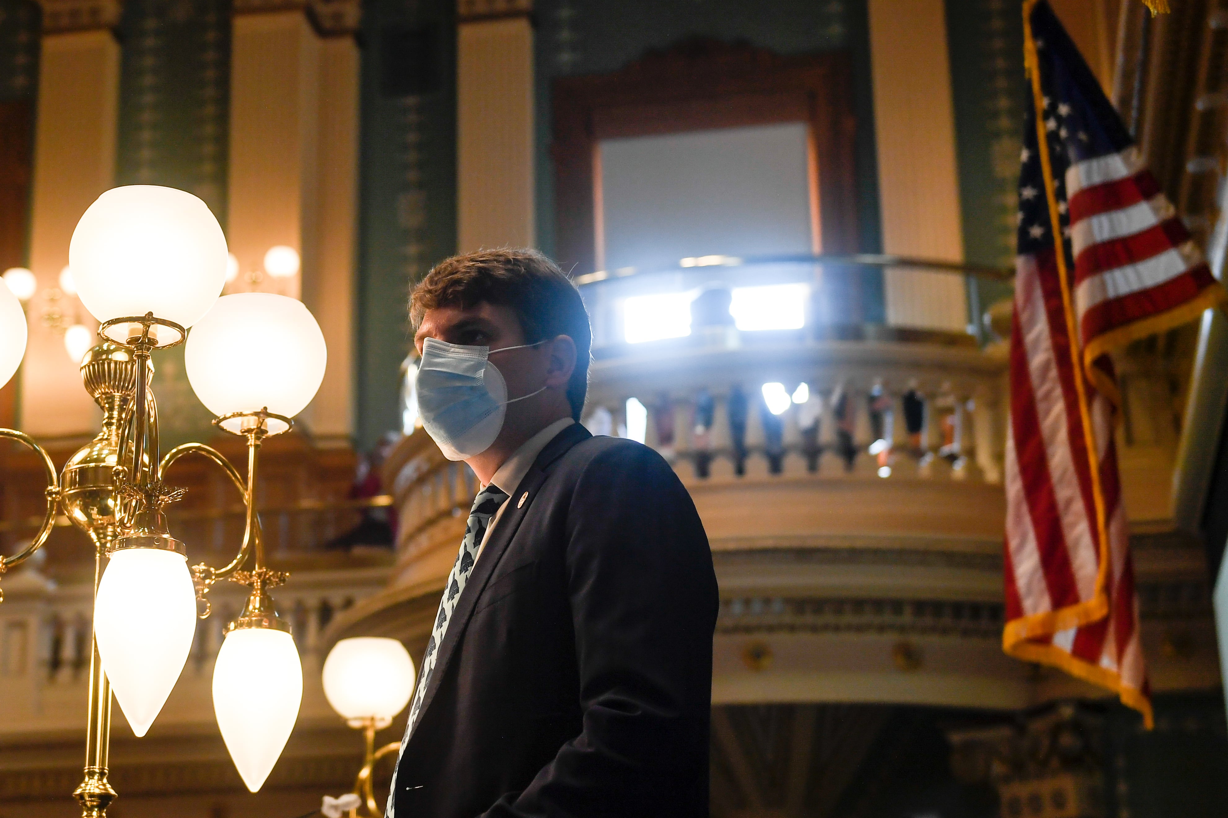 Colorado Speaker of the House Alec Garnett listens to an introduction in the State House chambers. He is wearing mask in an ornate room.