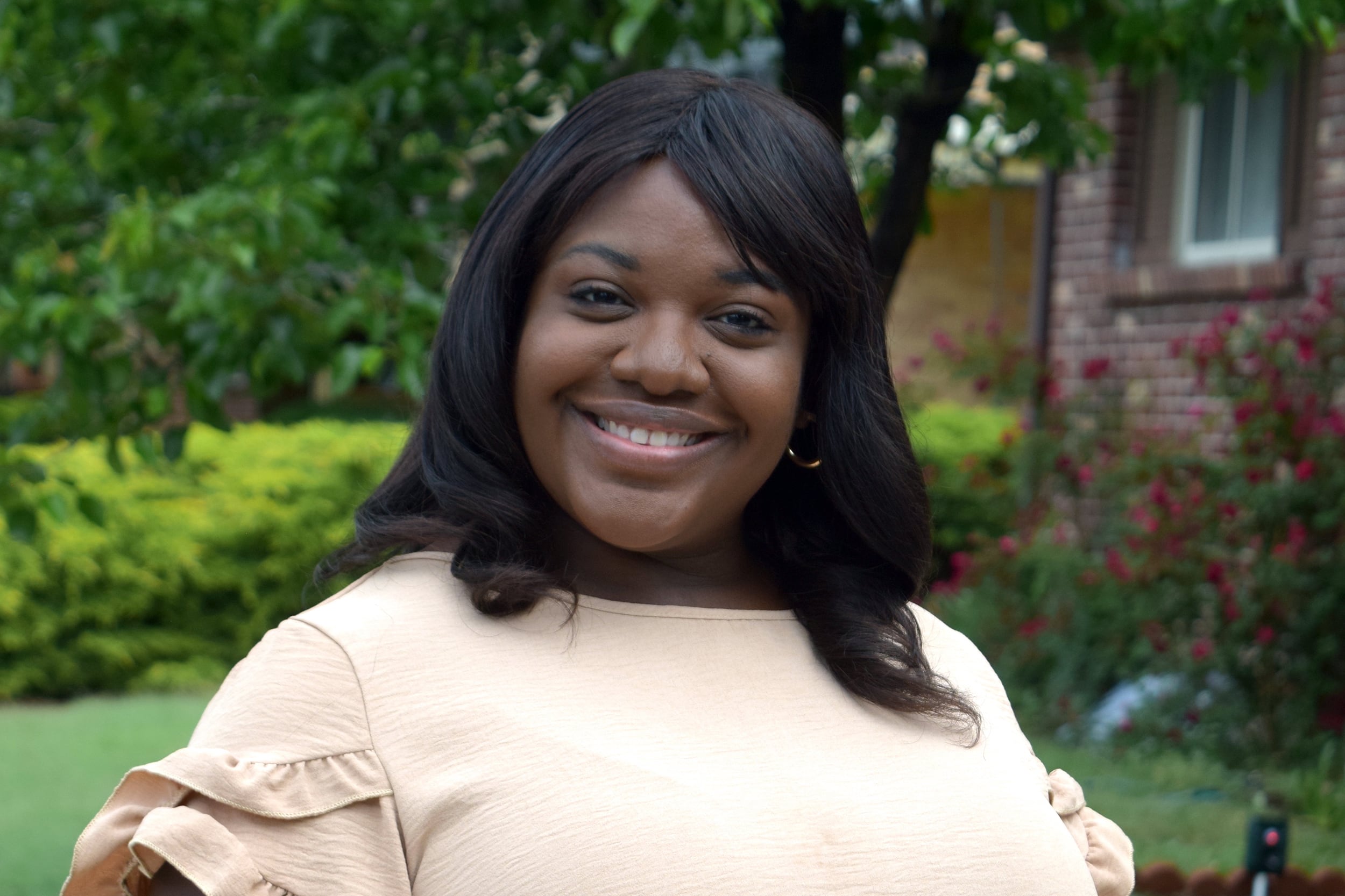 A photograph of a Black woman wearing a cream colored blouse with short dark hair poses for a portrait smiling outside with green foliage in the background.