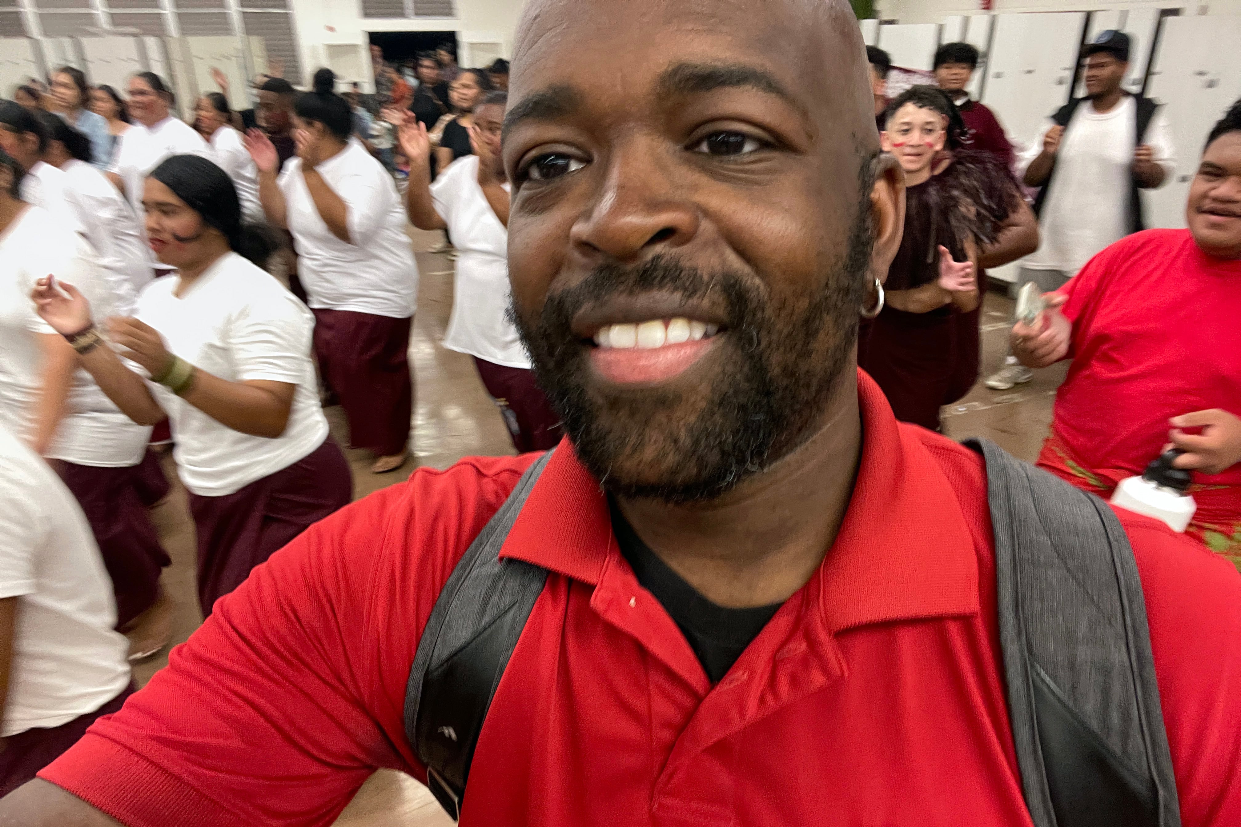 A close up of a man's face smiling at the camera and wearing a red shirt with students dancing in the background.