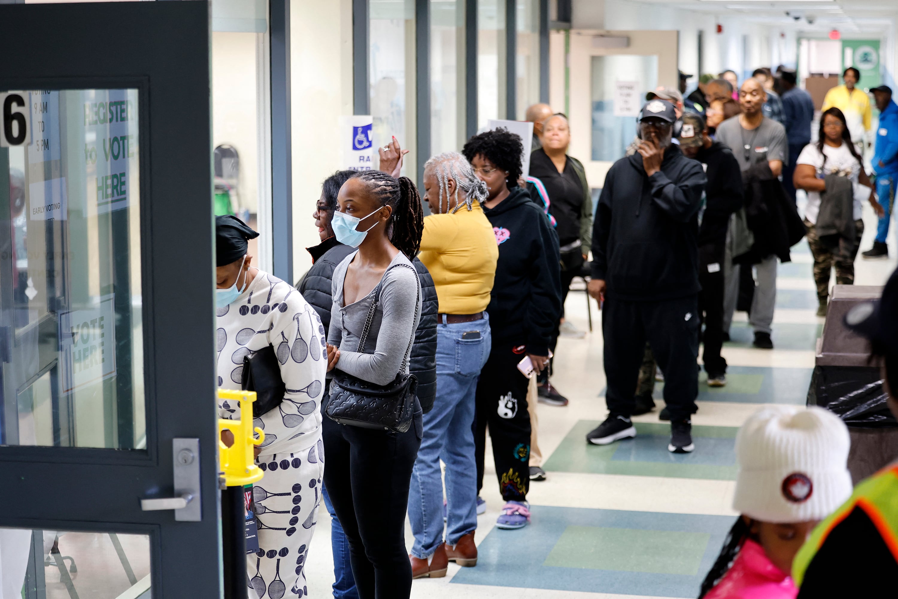 A line of people stand in front of a glass wall waiting to enter an early voting location in a hallway.