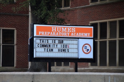 A white and orange sign that reads "Humes Preparatory Academy: This is our community 100% Team Humes" sits outside of a brick building.