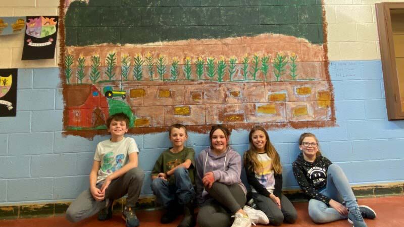 Five elementary children sit beneath a wall mural of mountains, a red sky with yellow sun, and a brown field.