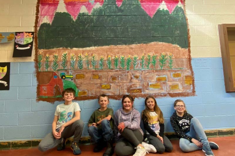 Five elementary children sit beneath a wall mural of mountains, a red sky with yellow sun, and a brown field.