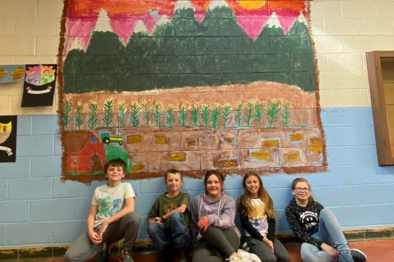 Five elementary children sit beneath a wall mural of mountains, a red sky with yellow sun, and a brown field.