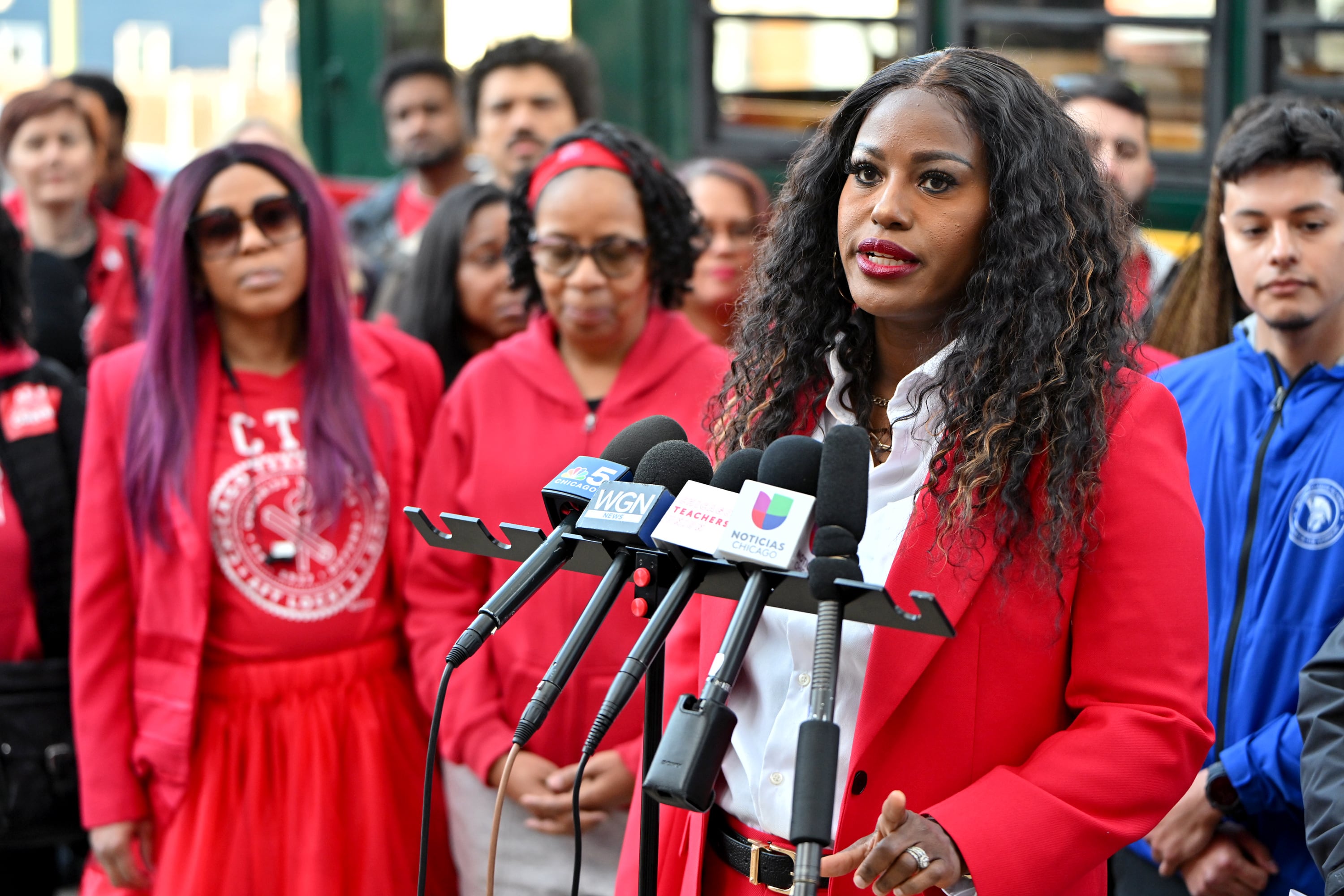 A person with short hair and wearing a red jacket, stand in front of microphones and speaks during a press conference with a group of people wearing red in the background.