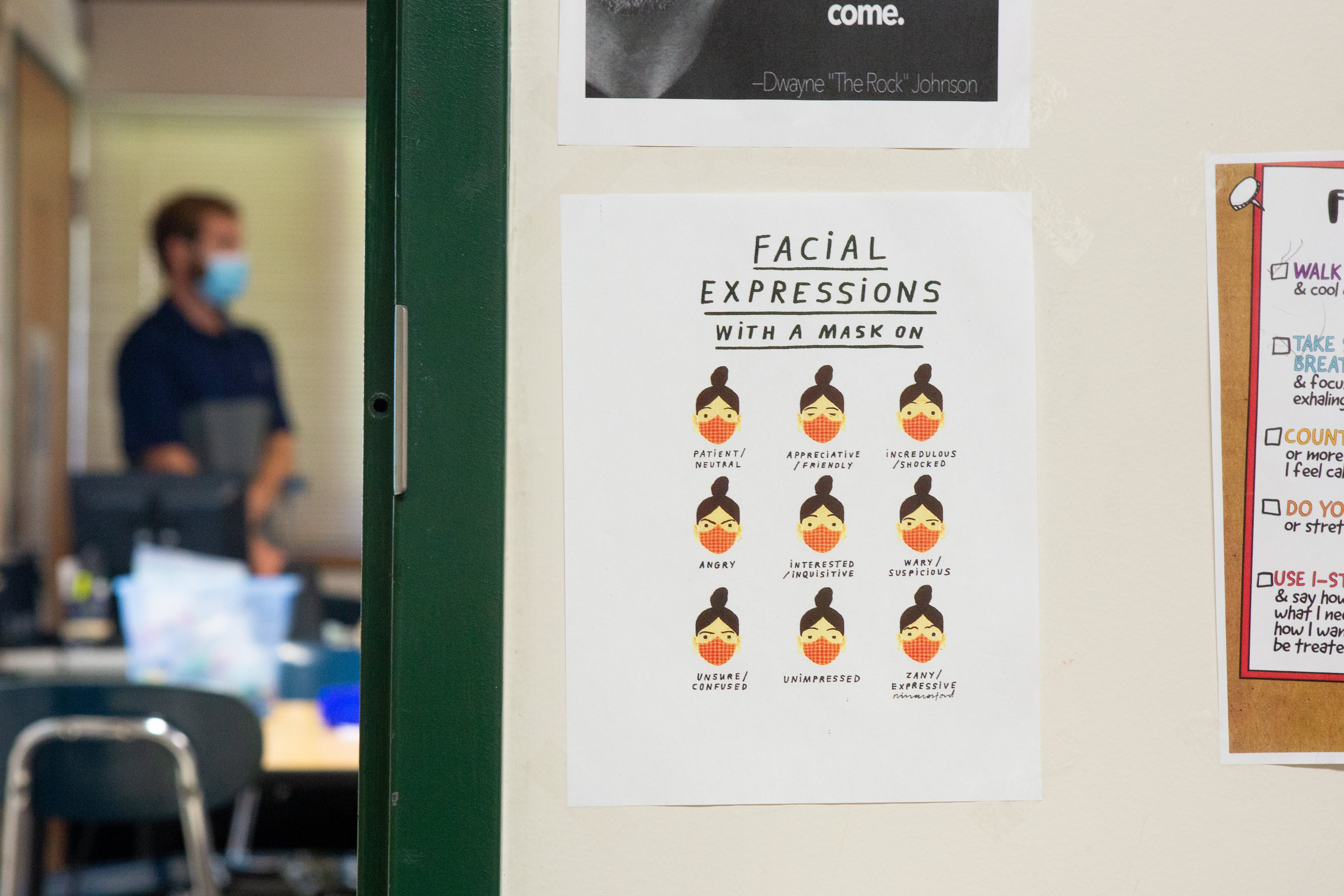 A sign that reads: Facial expressions with a mask on, posted on a wall outside a classroom, where a male is seen standing and wearing a mask.