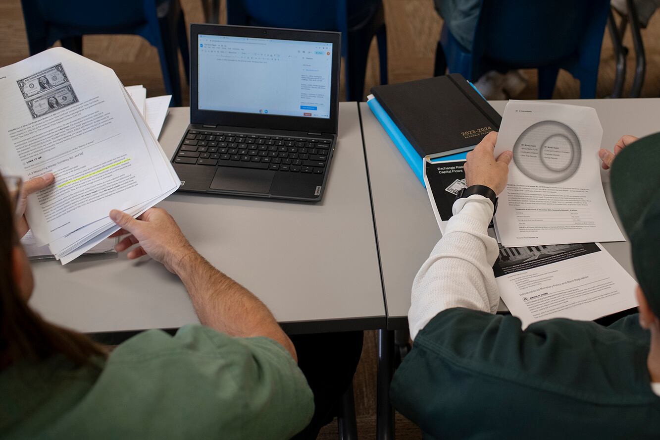 Two people sit at a table with papers and a computer in front of them. You can only see their arms and hands.