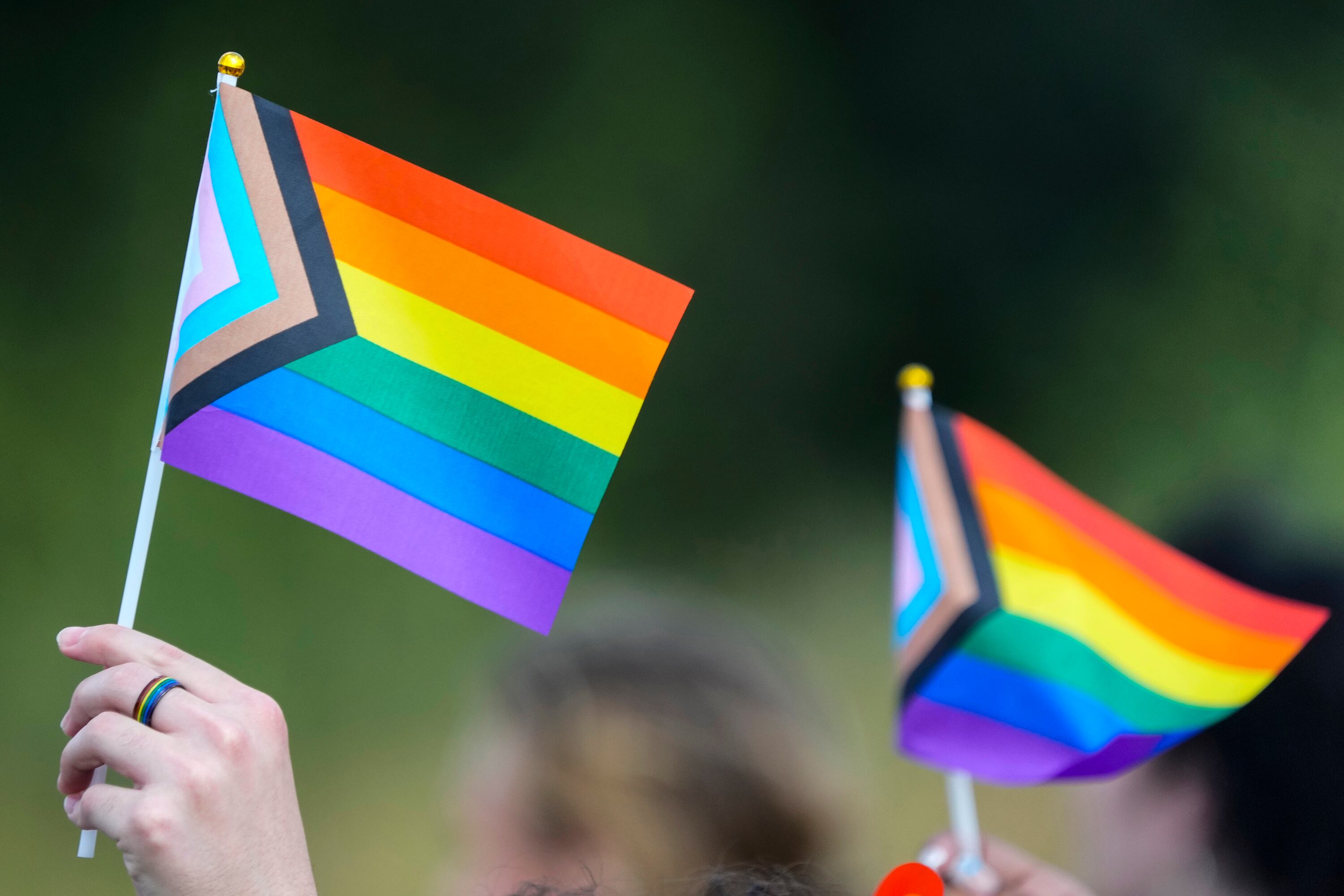 A close up of a hand in the air holding a pride flag with a small pride flag in the background.