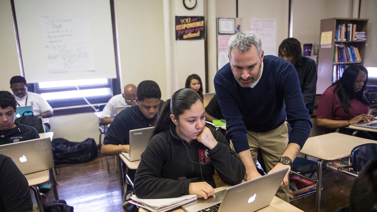 A teacher works with one student sitting at a wooden desk in a classroom full of desks in a classroom.