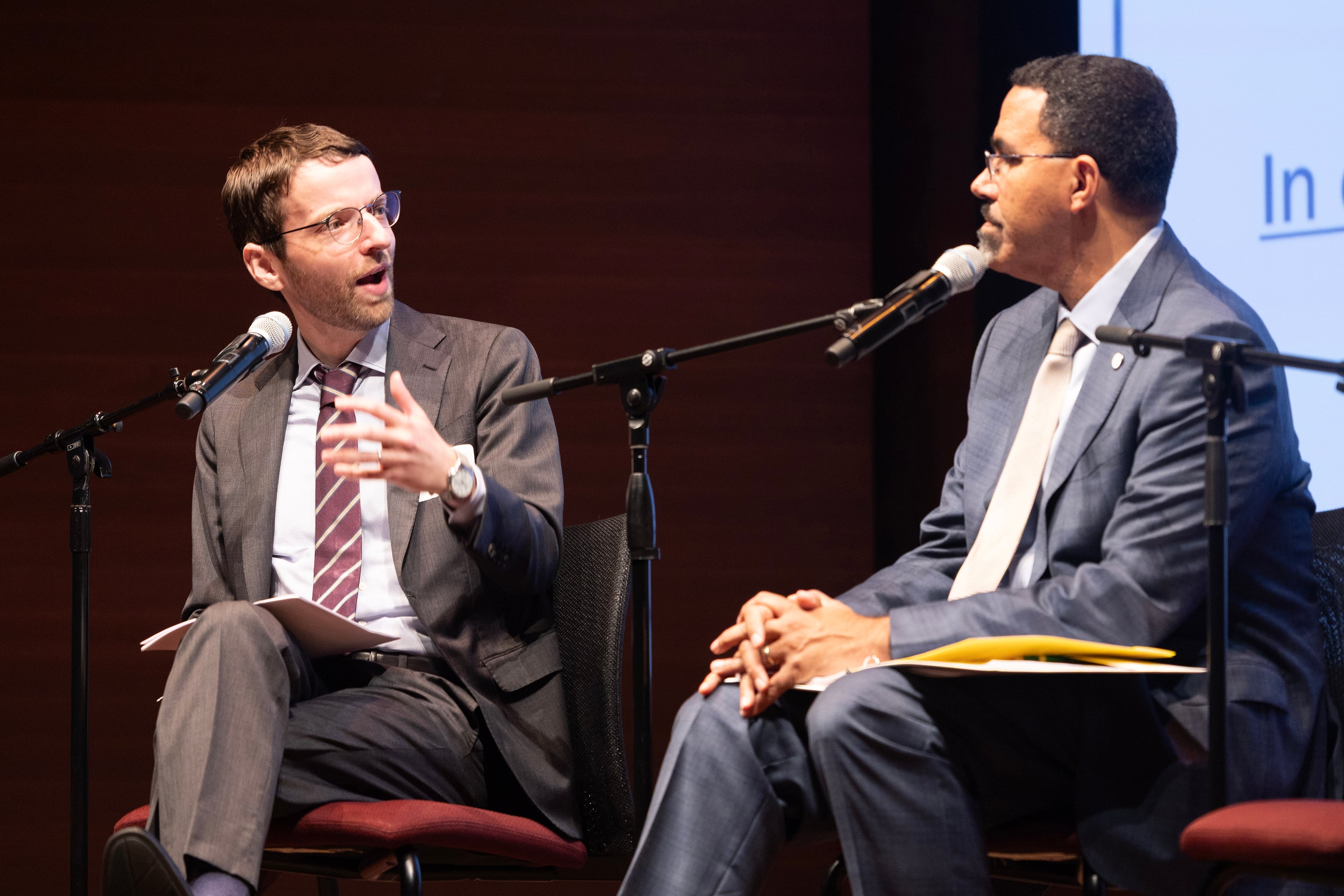 A photograph of two men in suits sitting in chairs next to each other on stage.