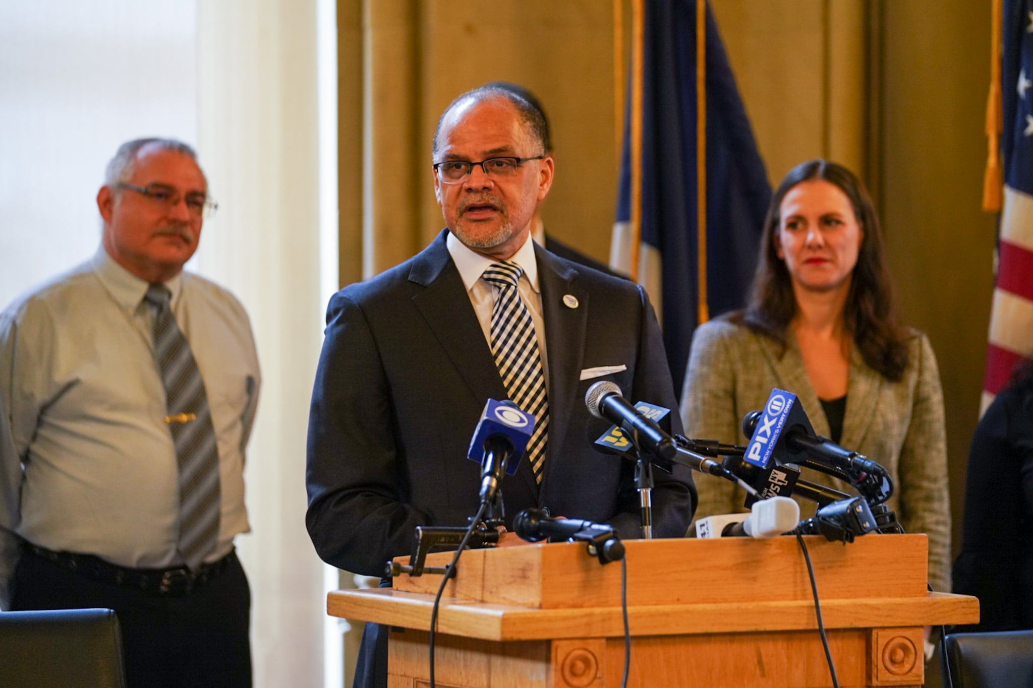 A man wearing a dark suit stands at a podium while two people stand behind him.