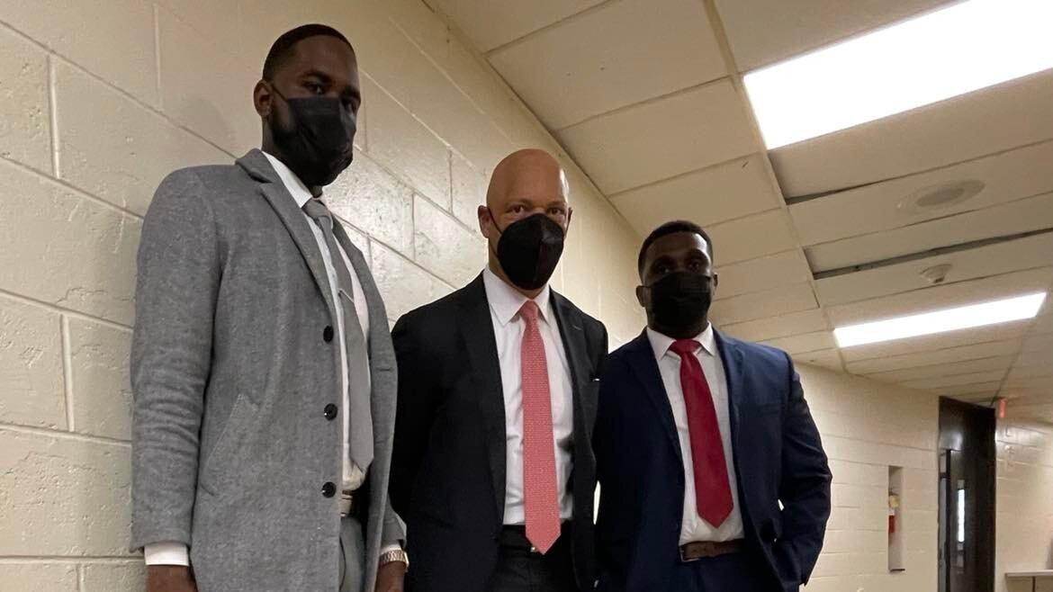 Three men is suits and wearing face masks stand together in a school hallway.