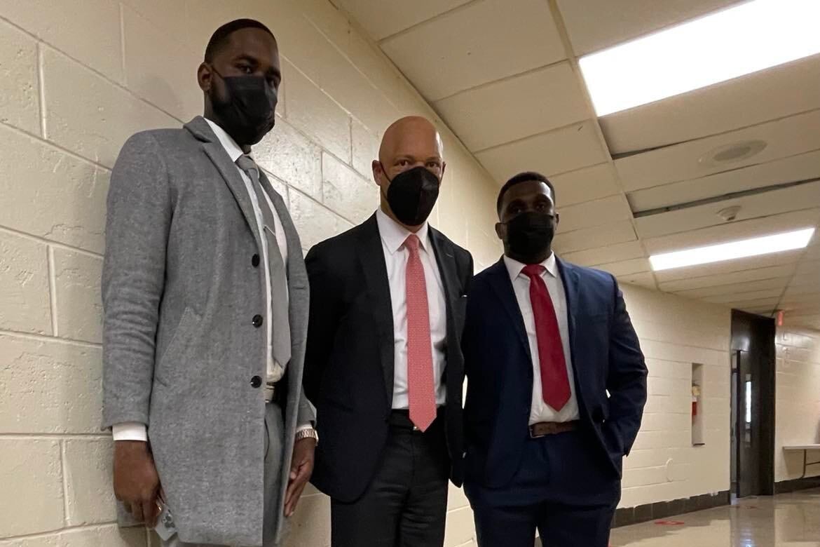 Three men is suits and wearing face masks stand together in a school hallway.