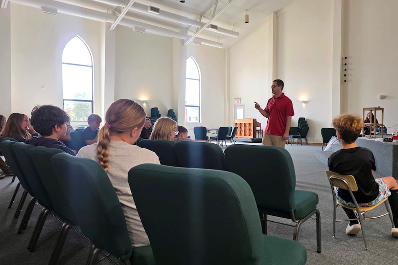 Students sit in rows of chairs inside of a large building while a teacher speaks at the front of the class.