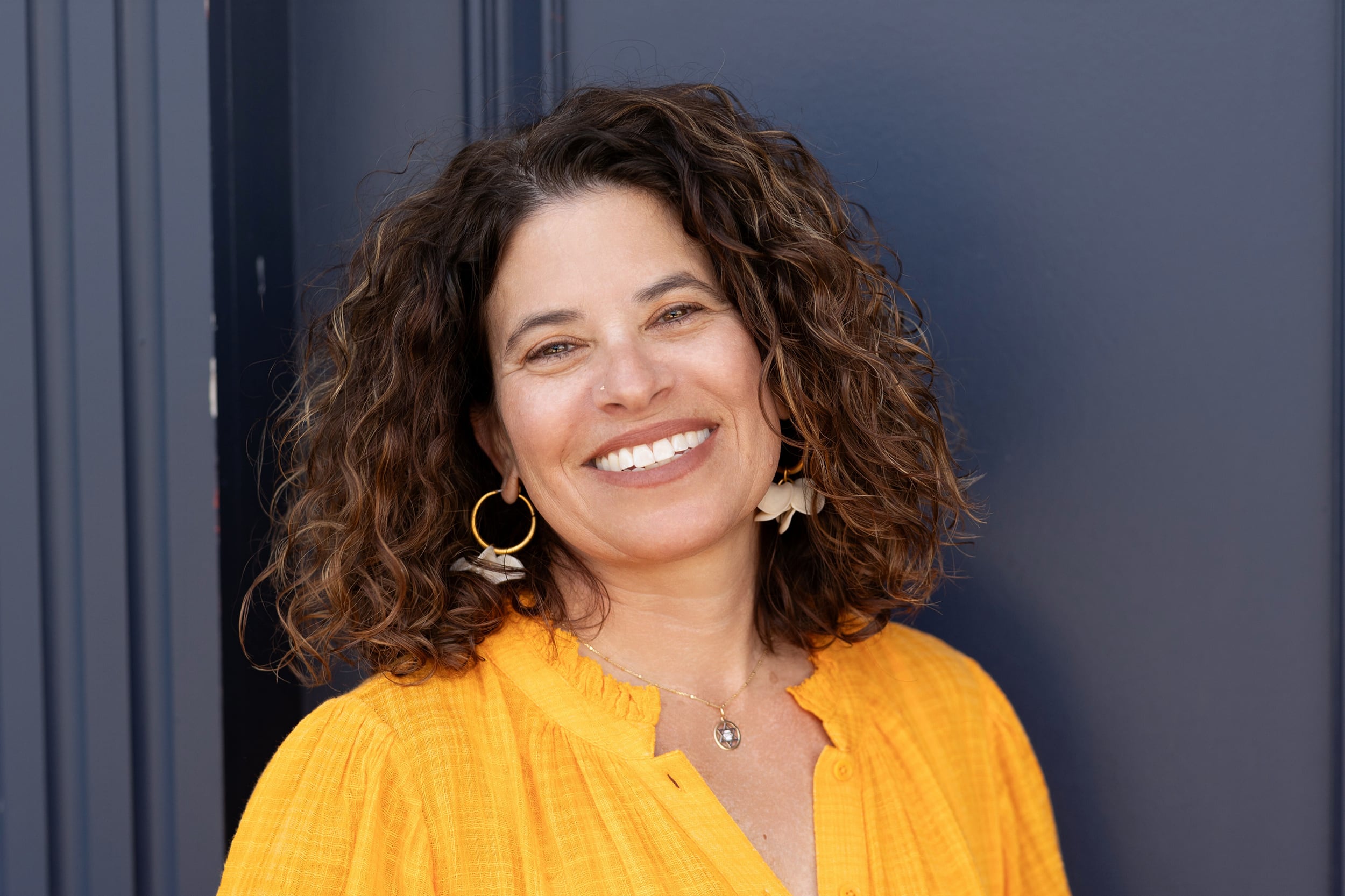 A woman with light skin, short dark, curly hair and wearing a yellow blouse poses for portrait in front of a grey background.