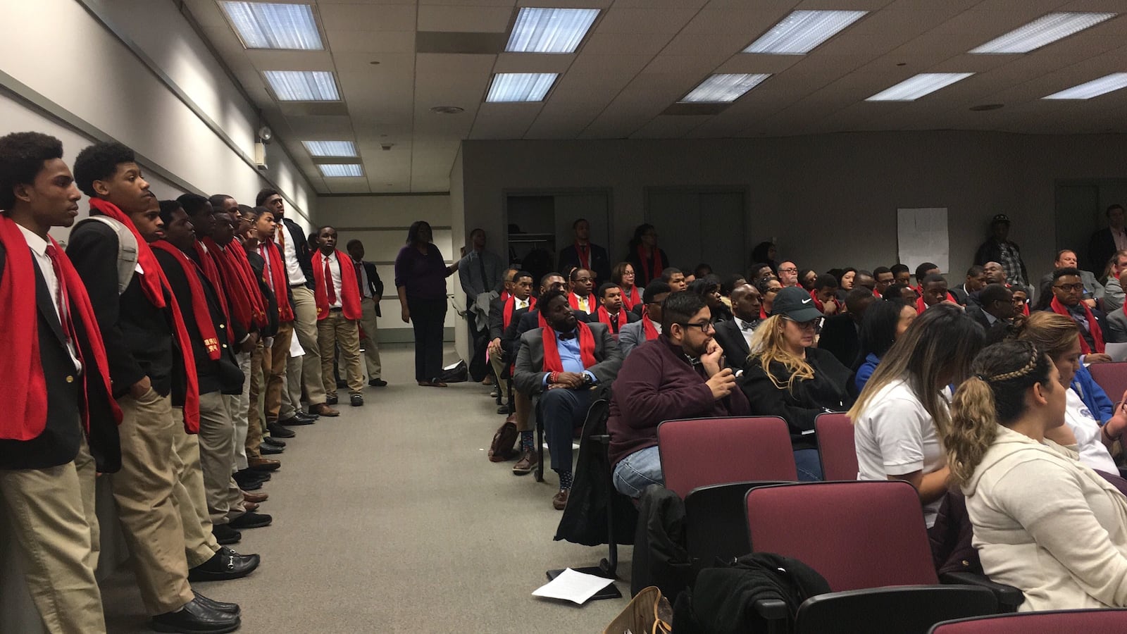 A group of high school students stand at left. Adults are seated nearby in an auditorium.