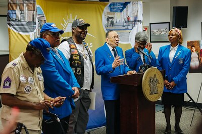 Six Black adults, some in suits and some in uniforms stand behind a podium with a microphone and in front of a yellow banner.