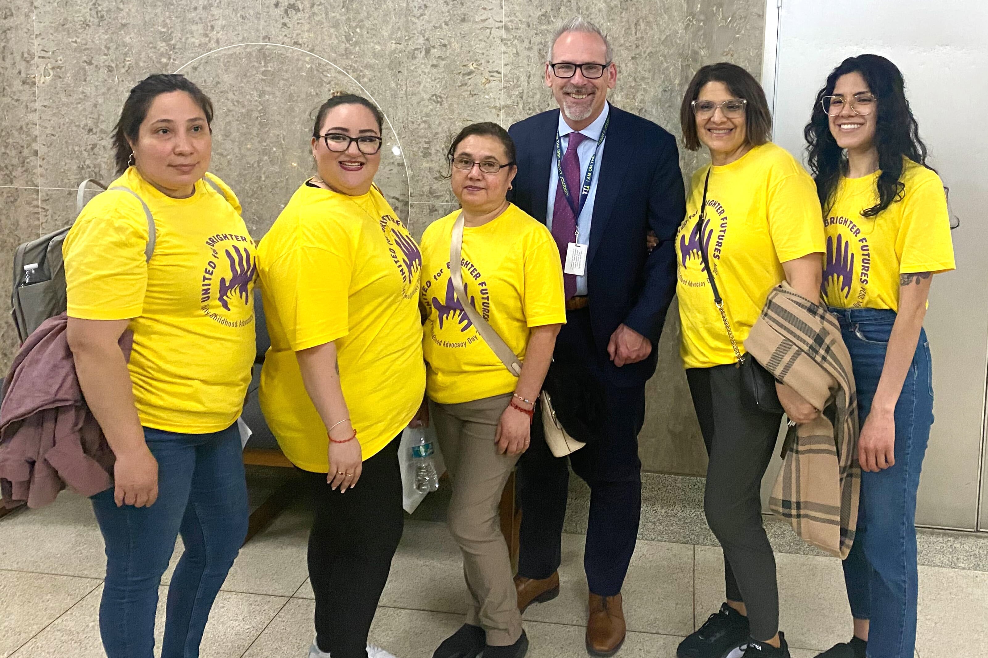 Five women in yellow t-shirts stand with a man in a suit.