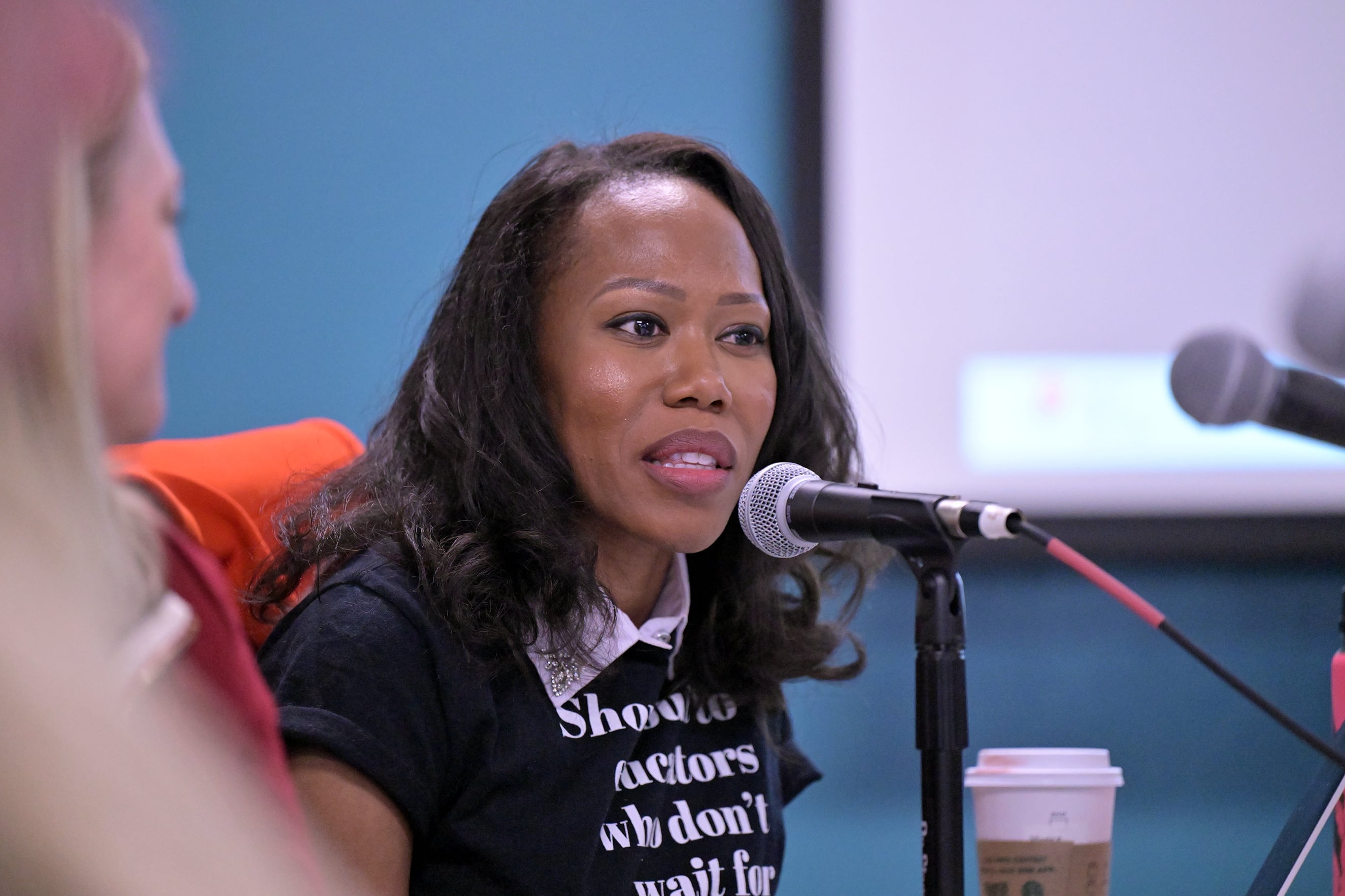 A Black woman with dark hair speaks from a microphone sitting in a conference room.
