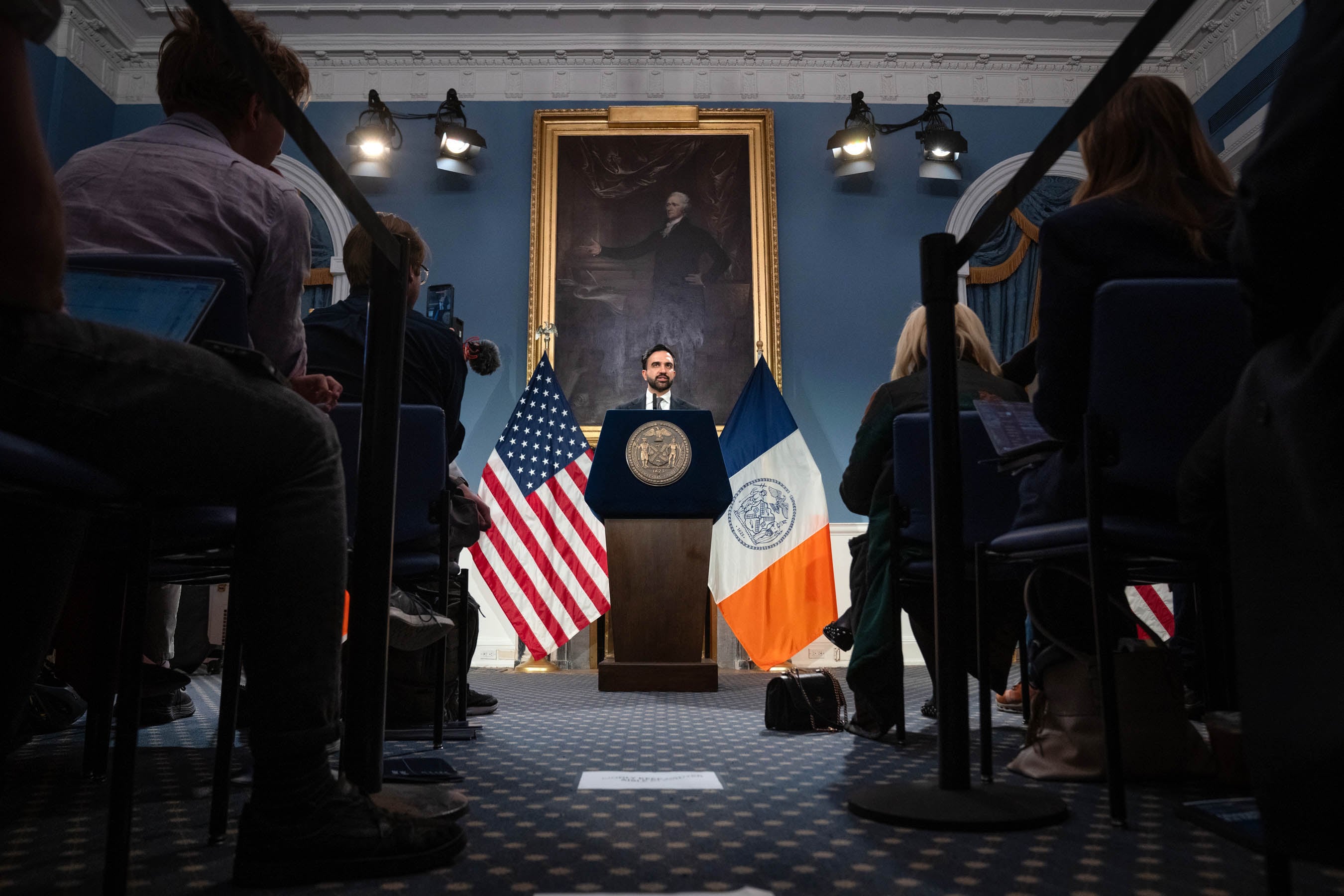 A photograph of a man in a suit standing behind a wooden podium speaking to a room full of people sitting in rows of chairs looking at him. Two flags are positioned behind the man in the center.