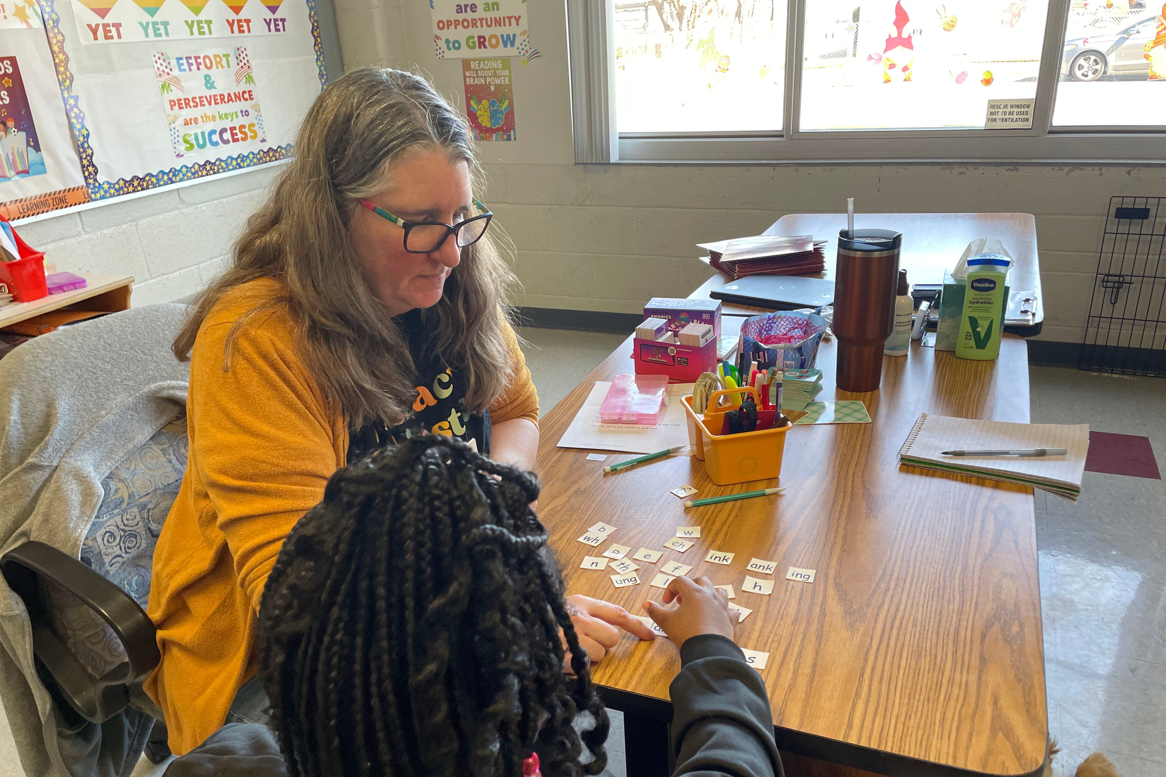 A white educator in a yellow sweater sits across the table from a young Black student with long, braided hair in a classroom.