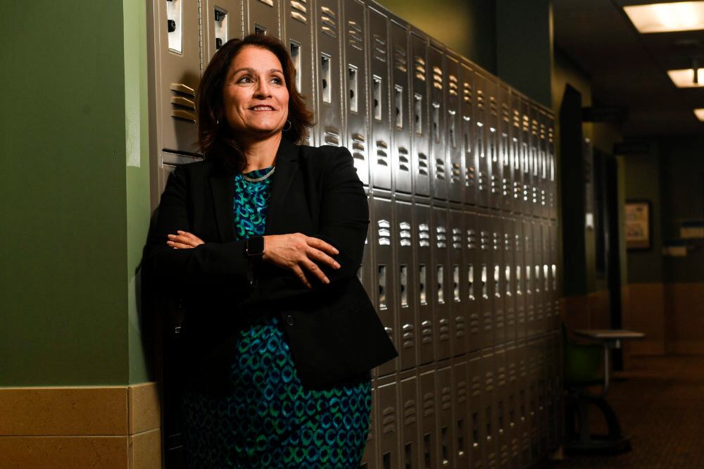 A woman leans against a wall of lockers. Her arms are crossed in front of her. She is wearing a black blazer.