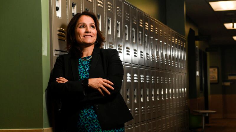 A woman leans against a wall of lockers. Her arms are crossed in front of her. She is wearing a black blazer.
