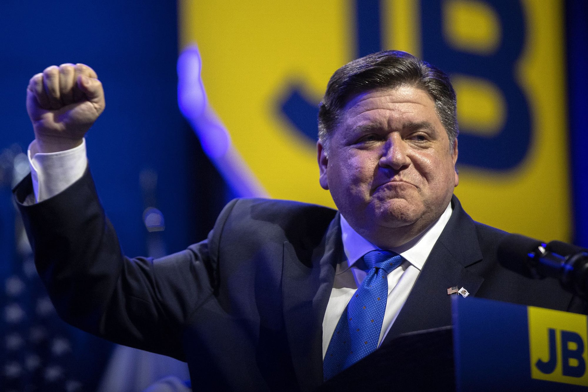 Gov. J.B. Pritzker stands at a podium with his fist raised in the area with a blue and yellow sign in the background with JB behind him