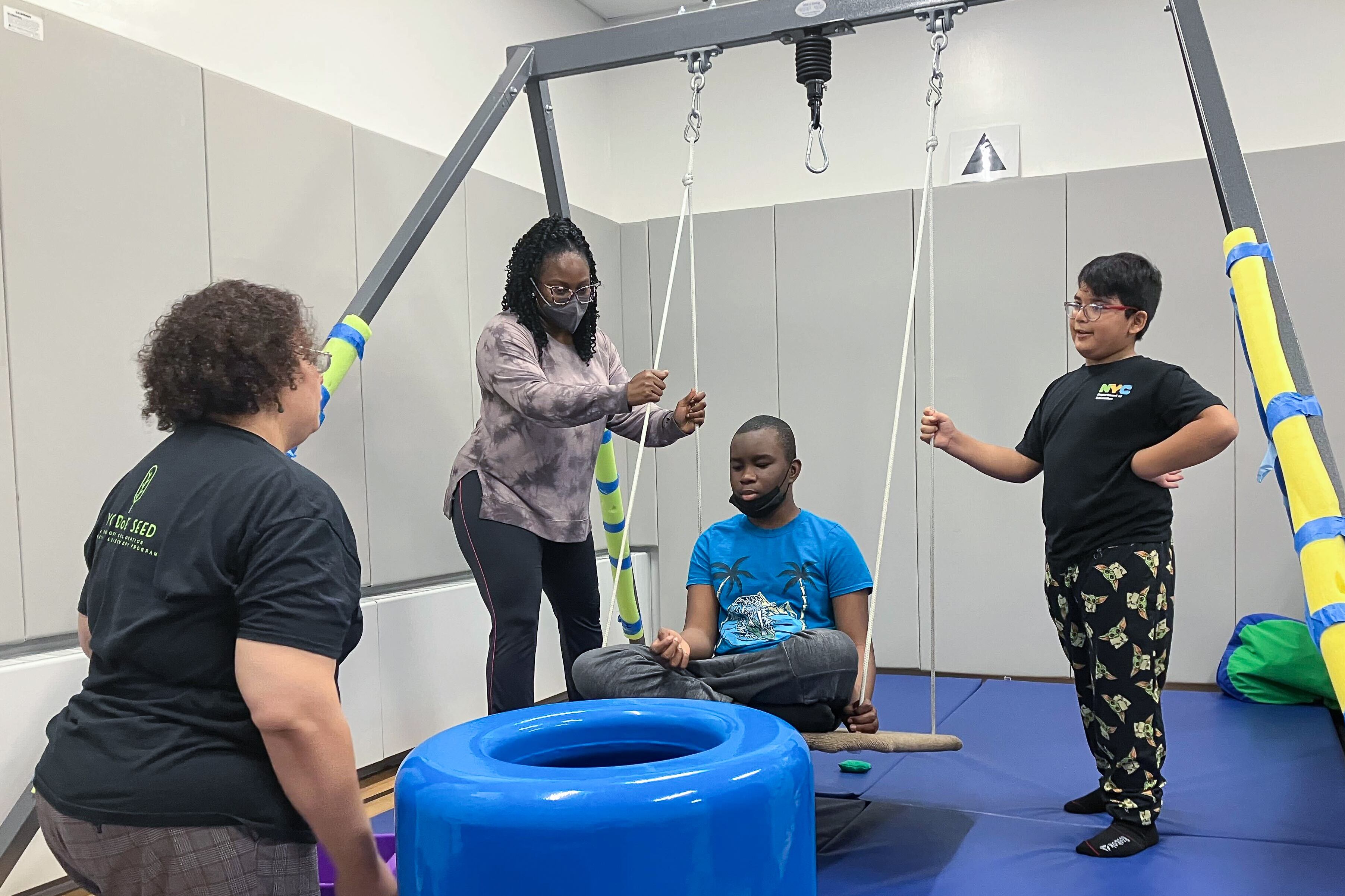 A child sits on a swing indoors with an adult and another child on either side.