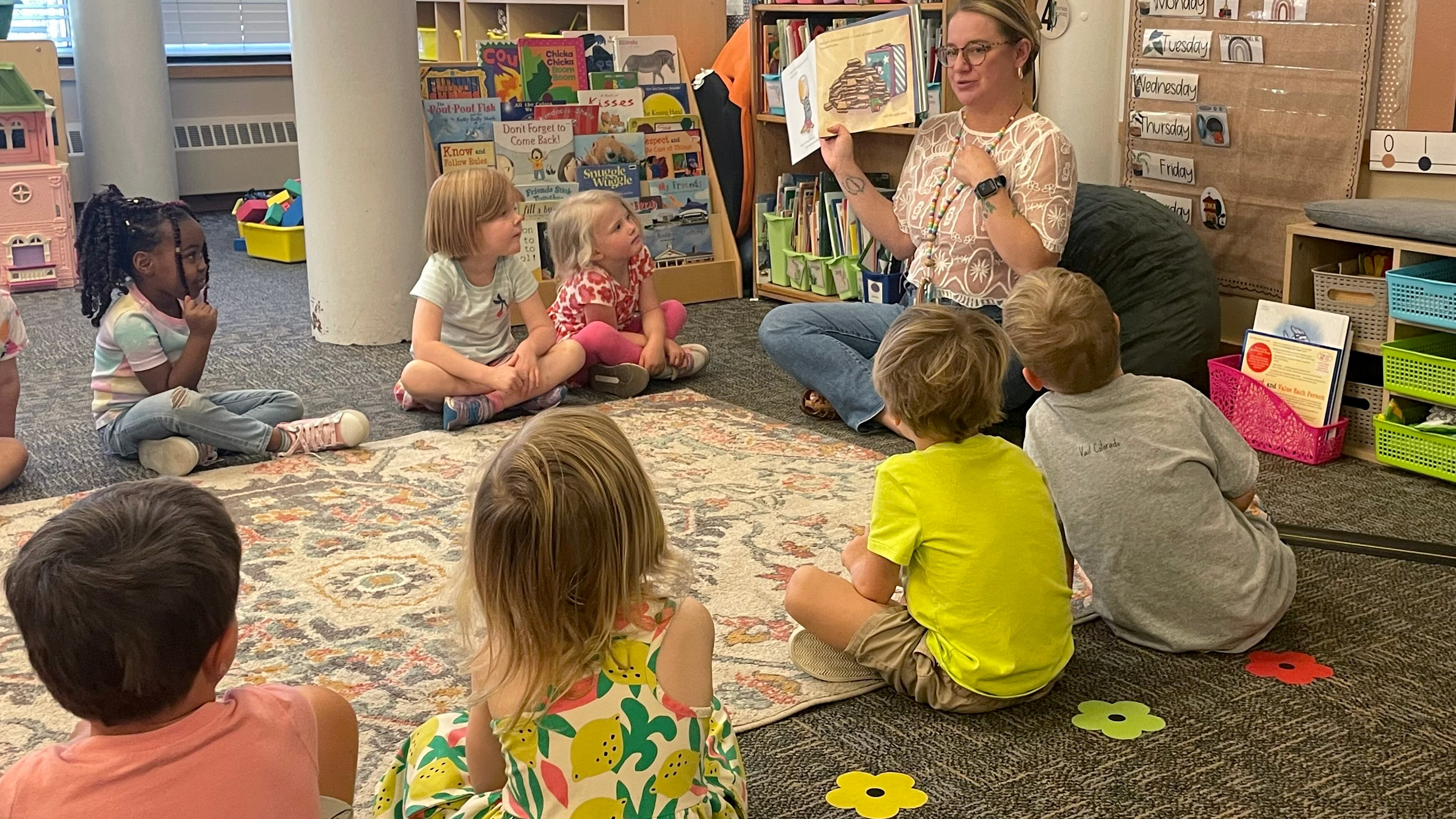 A teacher sitting cross-legged reads a picture book to preschool students sitting in a circle around her.