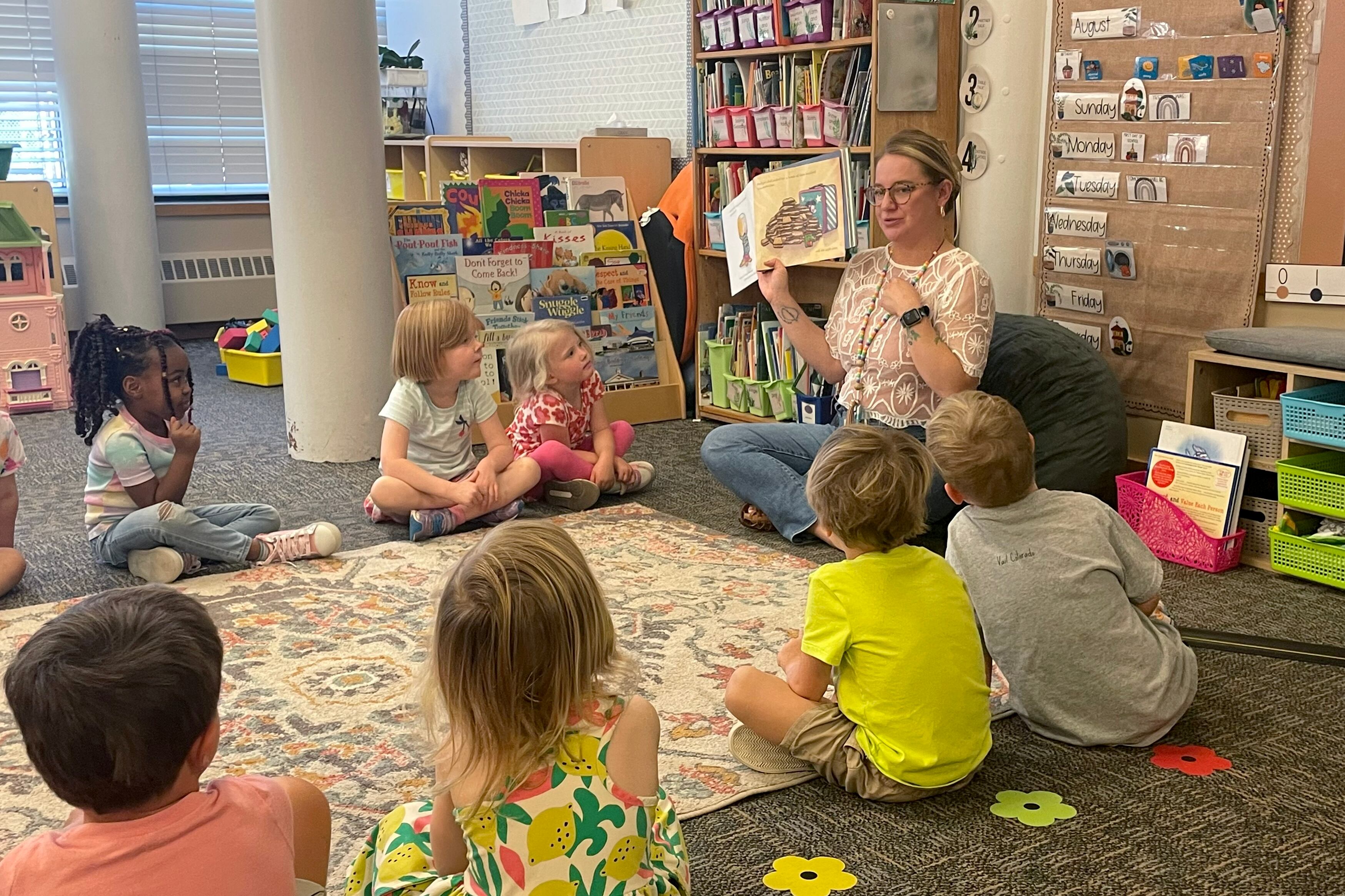 A teacher sitting cross-legged reads a picture book to preschool students sitting in a circle around her.