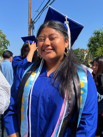 A high school graduate wearing a blue cap and gown with honor sashes poses for a portrait outside on a bright, sunny day.