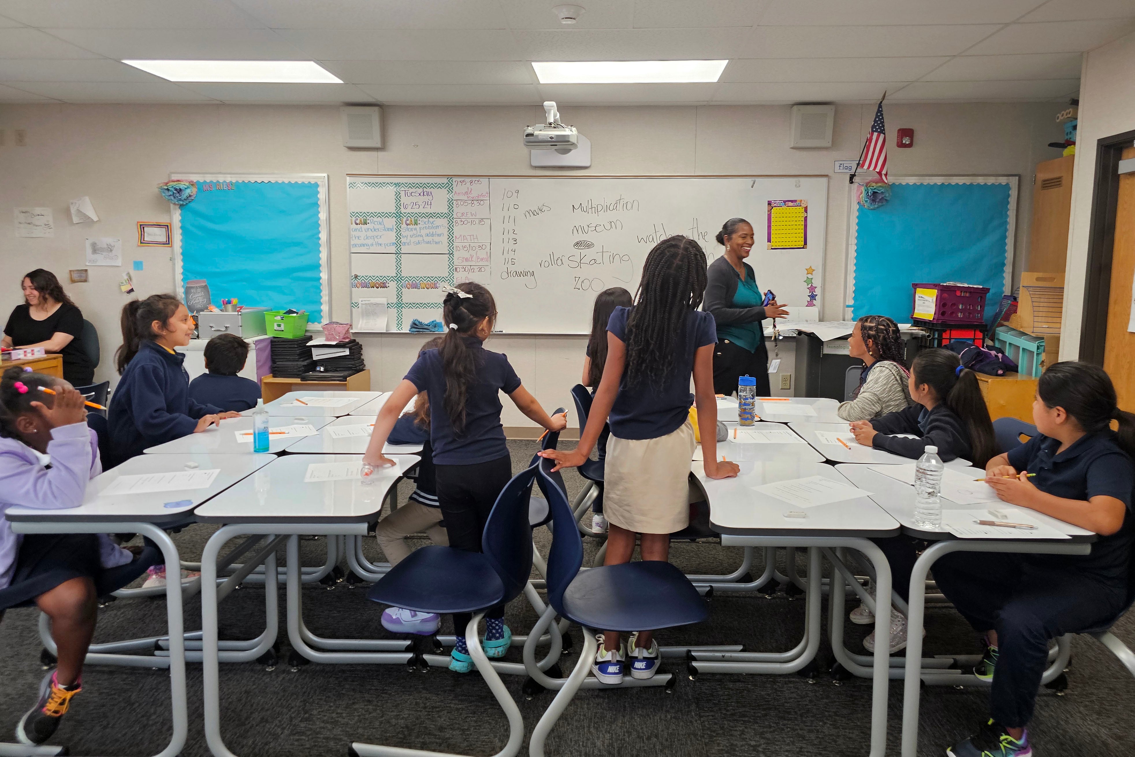 An adult stands toward the back of a classroom with students sitting at two tables.