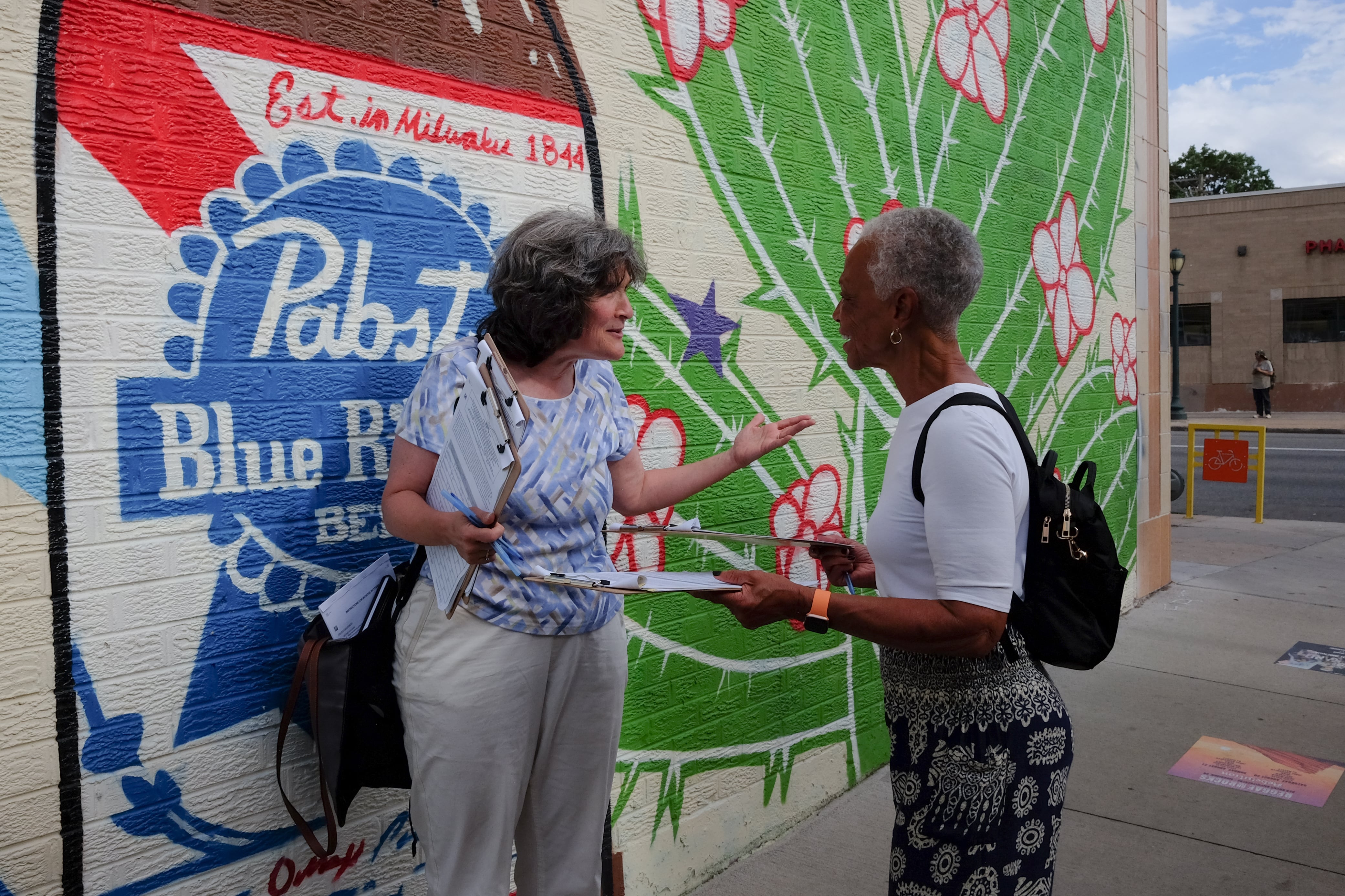 A white woman with shoulder-length salt-and-pepper hair and a blue-and-white shirt talks with a Black woman with short-cropped salt-and-pepper hair and a white t-shirt. Both women hold clipboards with signature sheets for a ballot measure. They’re standing against a wall with a mural that shows a cactus and a large Pabst Blue Ribbon bottle.