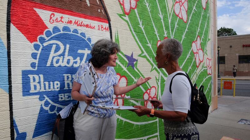 A white woman with shoulder-length salt-and-pepper hair and a blue-and-white shirt talks with a Black woman with short-cropped salt-and-pepper hair and a white t-shirt. Both women hold clipboards with signature sheets for a ballot measure. They’re standing against a wall with a mural that shows a cactus and a large Pabst Blue Ribbon bottle.