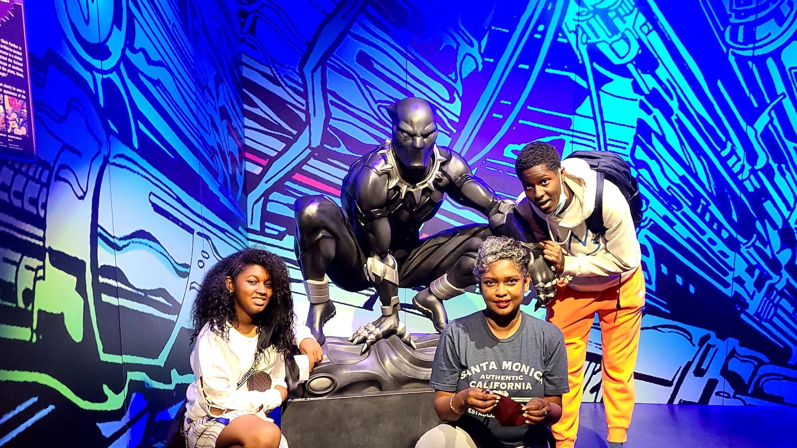 Two teens and an adult stand next to a statue. A colorful background is behind them.