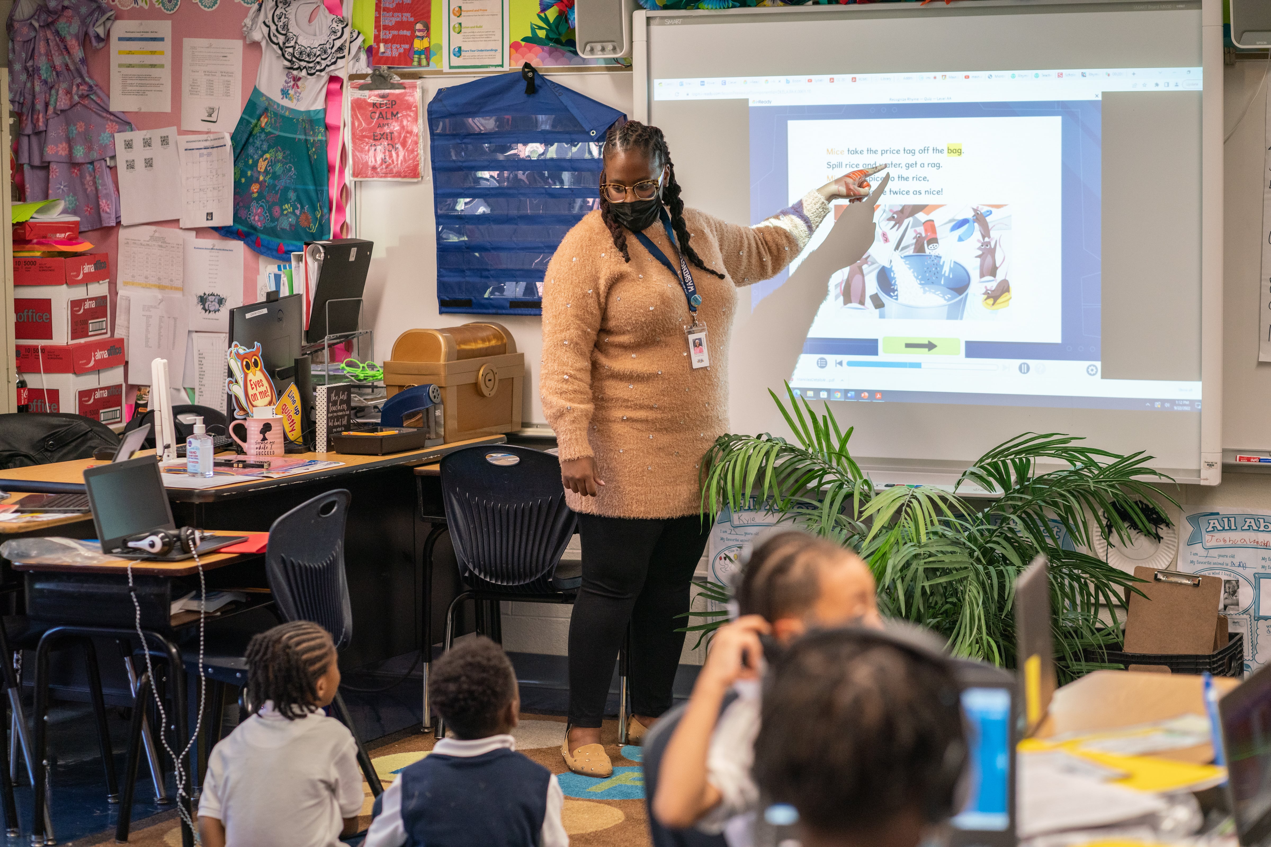 A woman points to a white board in the front of a classroom with students seated on both the floor and at desks.