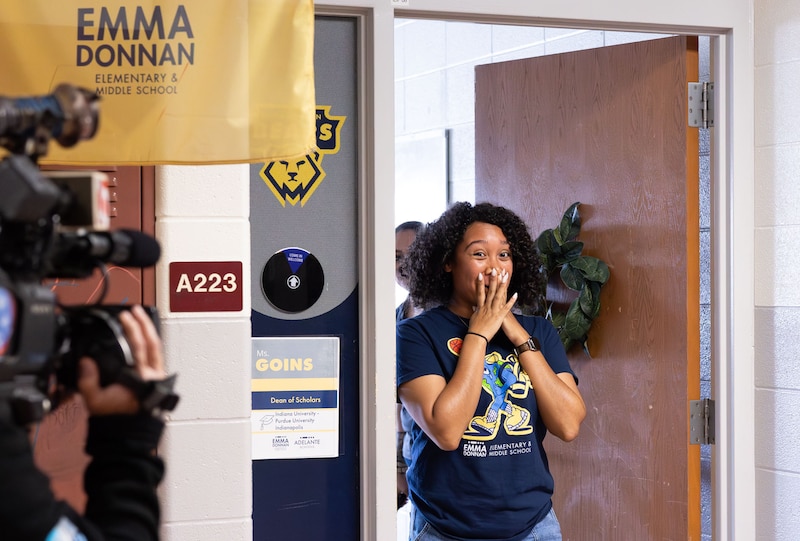 A woman with short dark hair and waring a dark blue shirt has her hands over her mouth as she is surprised by winning an award.