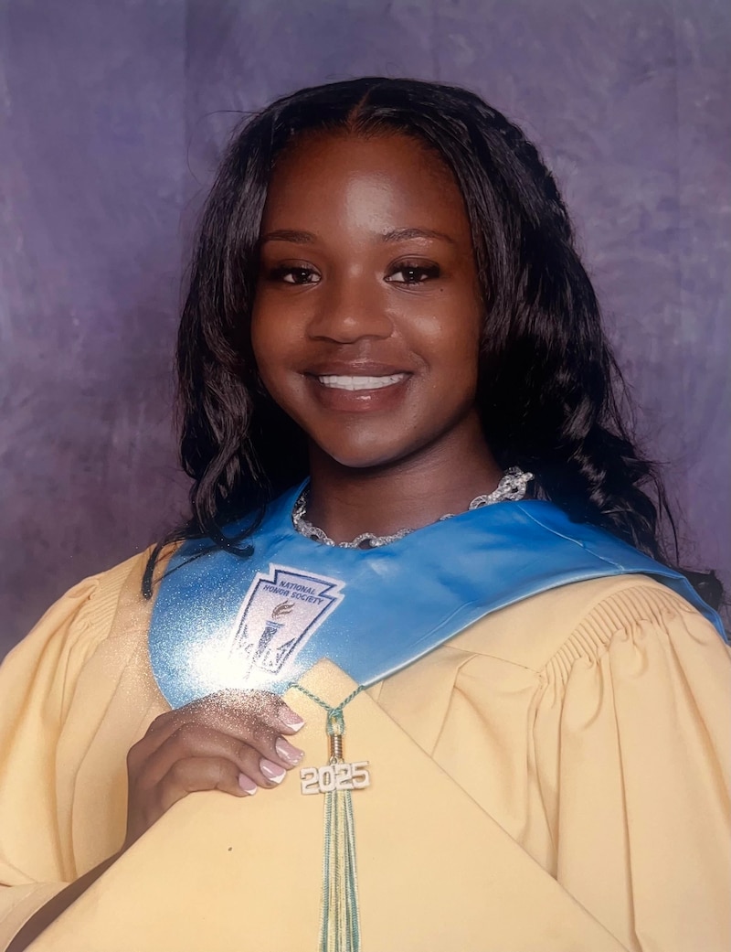 A Black high school senior girl with long dark hair and wearing a graduation gown and holding a graduation cap for a portrait.