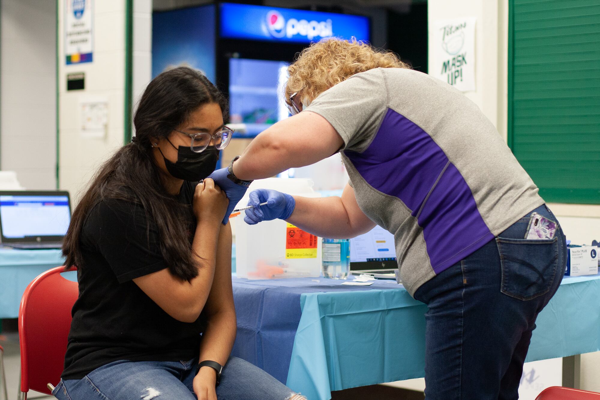 A young girl sitting on a red chair wearing a black shirt with a long, black ponytail holds up her sleeve while a woman in a blue and gray shirt stands next to her and inserts a syringe in her shoulder.