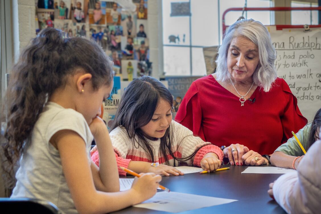 Two young female students work at a desk while a teacher wearing a red shirt with white hair looks on.