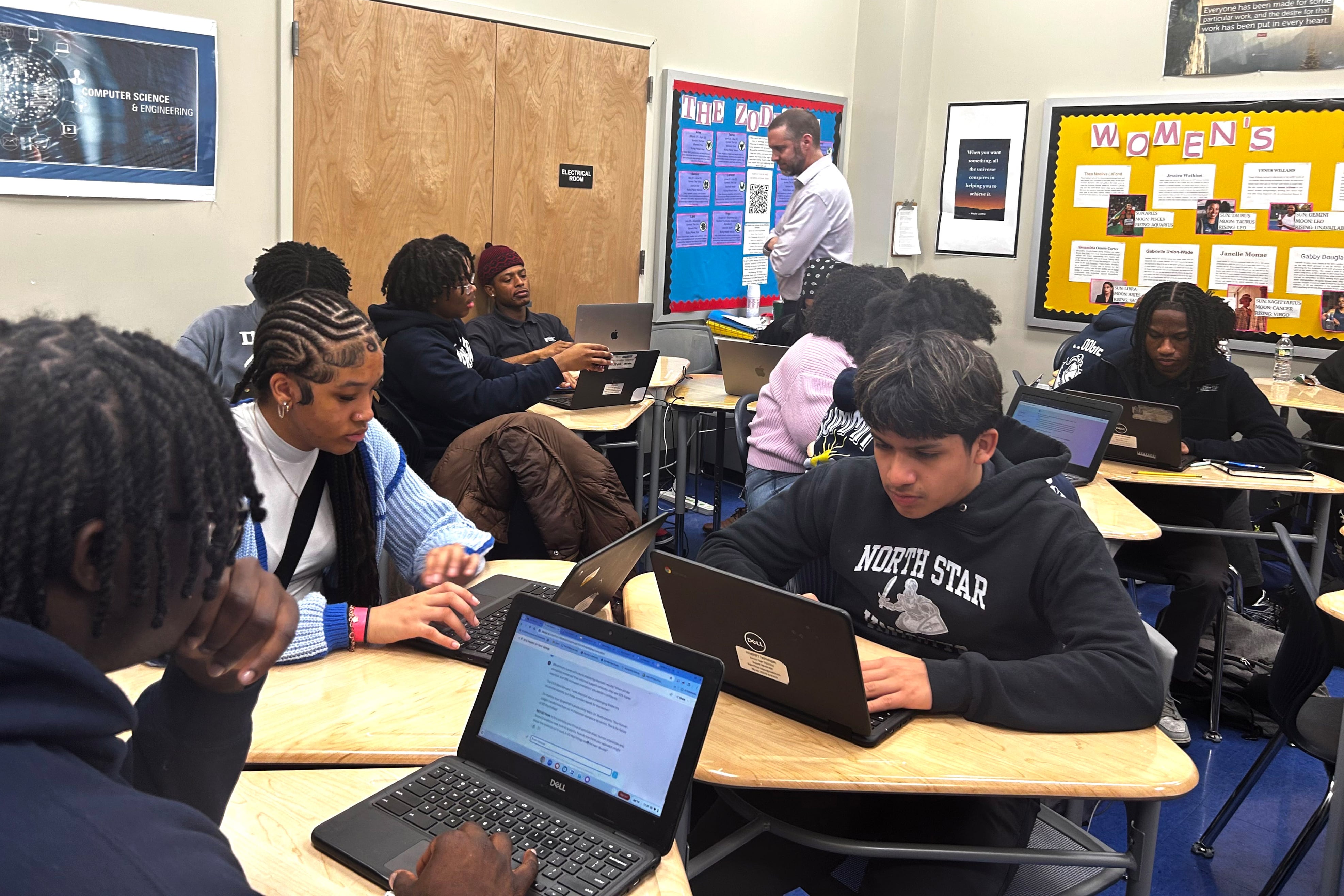 A man in a light shirt talks to a students while others work at tables.
