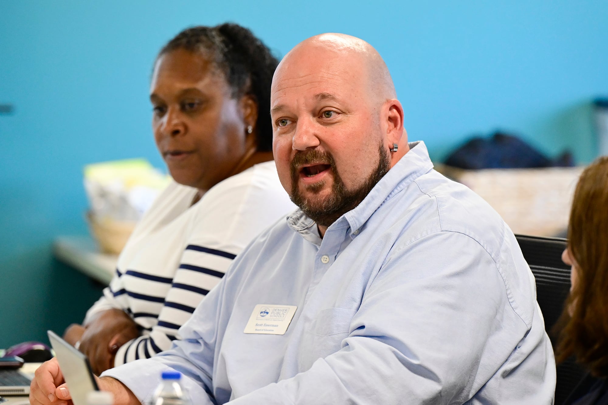 A white, bald man wearing a light blue dress shirt sits next to a Black women wearing a white and black striped sweater in a room with a blue wall in the background.