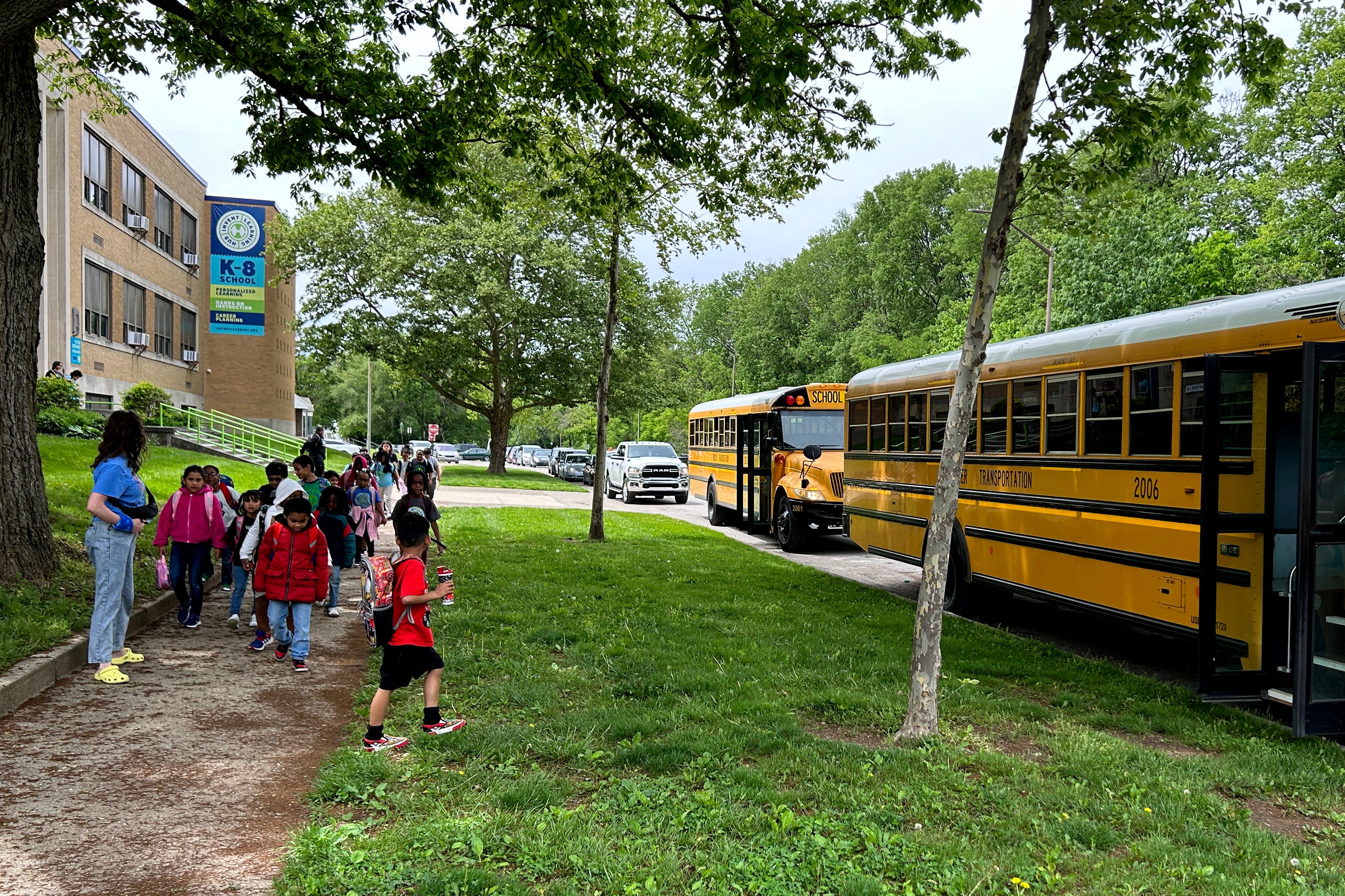 A group of young students line up outside with two yellow and black school busses near by.