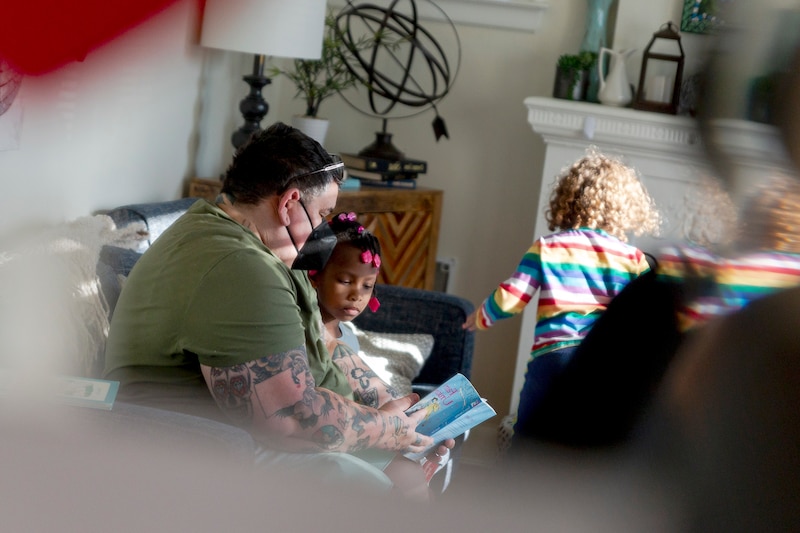 A person wearing a mask and a green shirt reads a book to a young child while sitting on a couch and others walk around them.